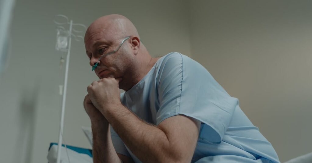 Bald man in hospital gown sits pensively with oxygen tube in healthcare setting.