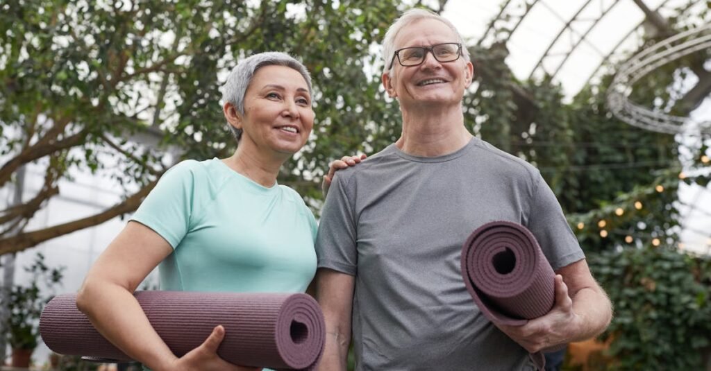 Happy senior couple holding yoga mats in a lush greenhouse setting.